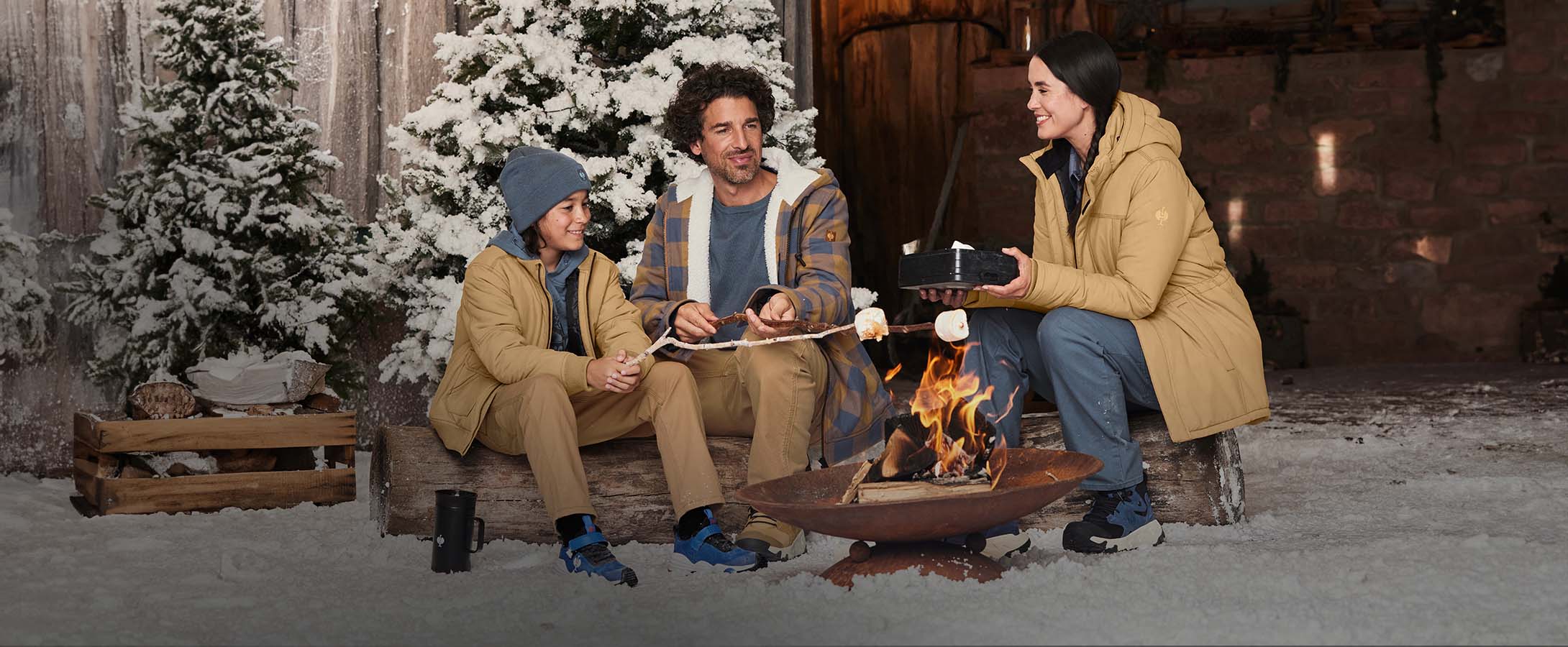 Woman, man, and boy wearing clothing from the e.s.iconic collection in a winter setting by a camp fire